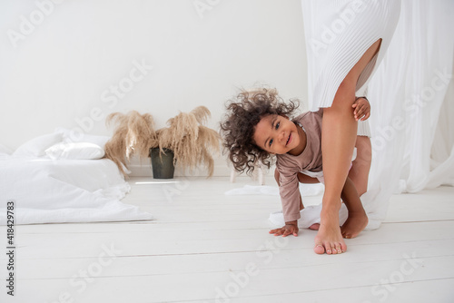 Little curly-haired African-American girl, hiding under mother's dress, peeking through legs, having fun, laughing, showing her tongue. Minimalist interior, pampas grass, white bed. Scandinavian style