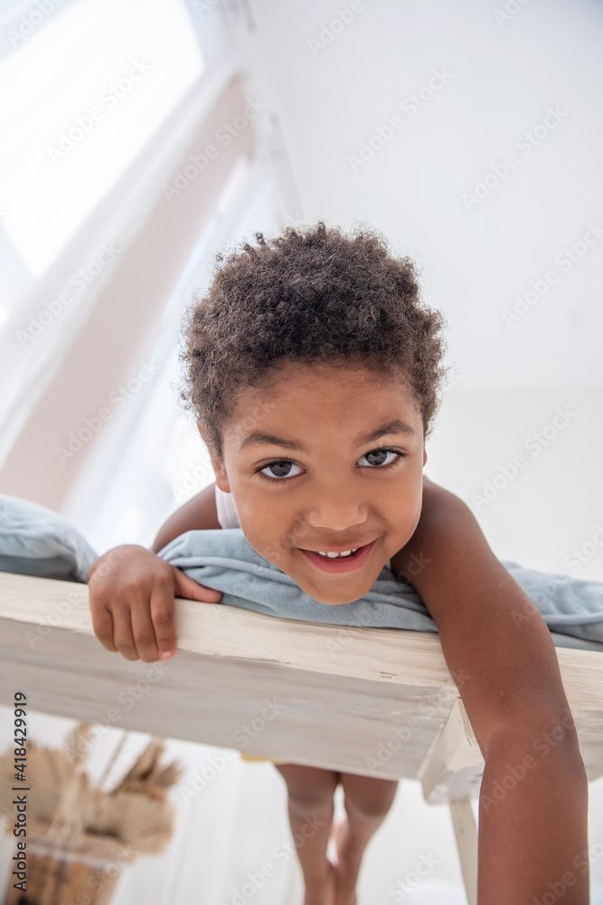 Fototapeta premium Little African American boy has fun, lies on white wooden chair, flounders with his feet. Minimalist interior, pampas grass, driftwood on the wall, airy doors on the windows. Scandinavian style.