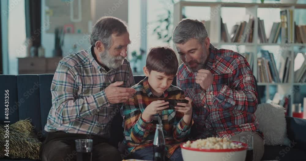 Young teenager boy playing computer game mobil ephone getting much support from his father and grandpa spending leisure time together.