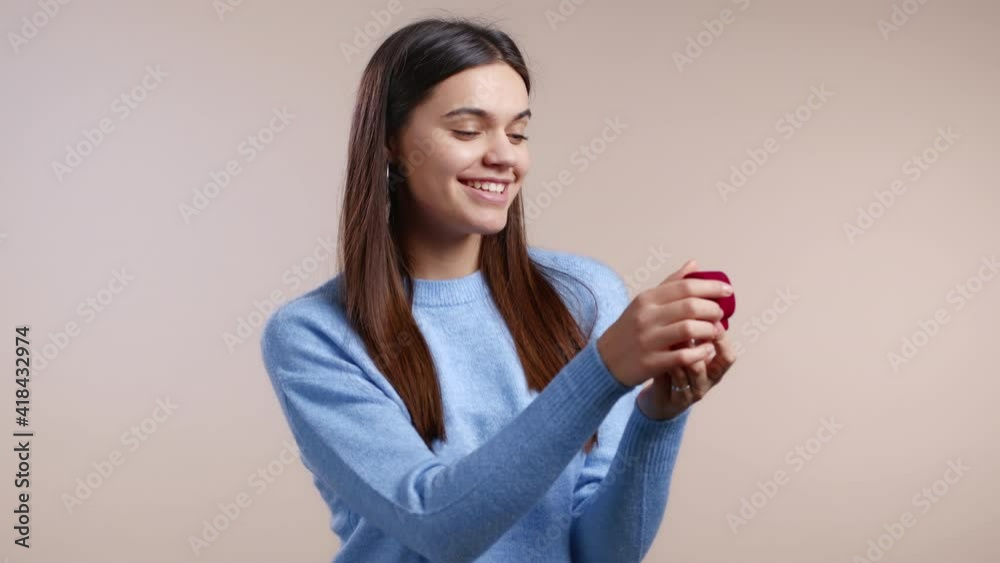  Pretty european woman in blue holding small jewelry box with marriage proposal on light studio background. Disappointed upset girl, she doesn't like gift.