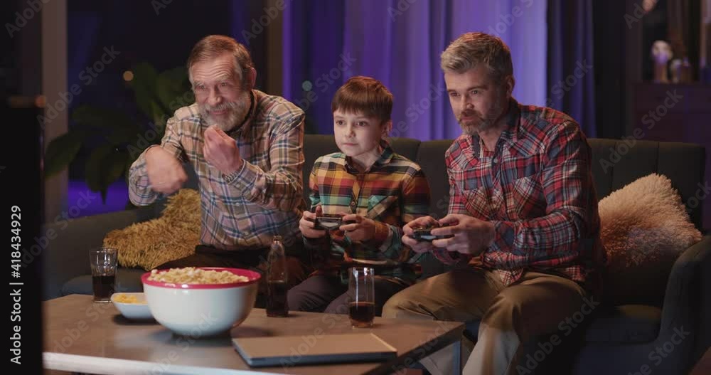 Home leisure. Family concept. Grandfather with his son and grandchild playing computer console video games with joysticks in cozy living room. Home leisure. Family concept. Grandfather with his son and grandchild playing computer console video games with joysticks in cozy living room.