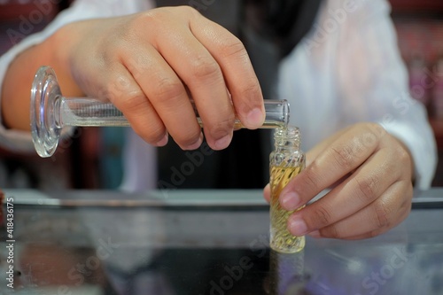 The hand of a perfume seller is pouring refillable perfume from a measuring cup into a small container