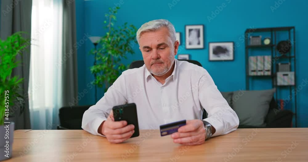 Mature 50s man sit at table use smart phone makes purchase on internet e-commerce retail websites, enters credit card data pay use e-bank application smiles enjoy easy fast money transfer remotely Mature 50s man sit at table use smart phone makes purchase on internet e-commerce retail websites, enters credit card data pay use e-bank application smiles enjoy easy fast money transfer remotely