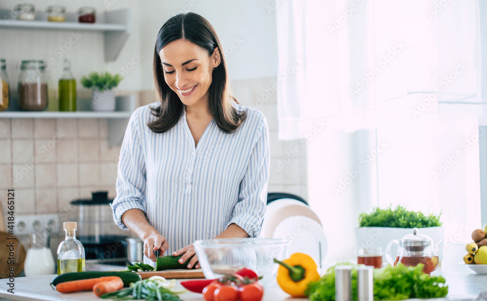 Cute happy young brunette woman in good mood preparing a fresh vegan salad for a healthy life in the kitchen of her home