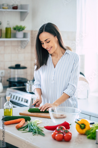 Cute happy young brunette woman in good mood preparing a fresh vegan salad for a healthy life in the kitchen of her home