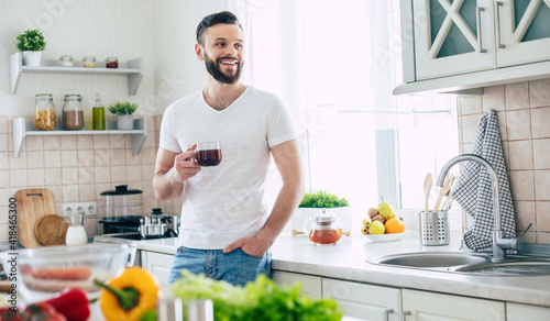 Handsome modern smiling young bearded man drinks some coffee or tea from his cup and relaxing in the kitchen at home