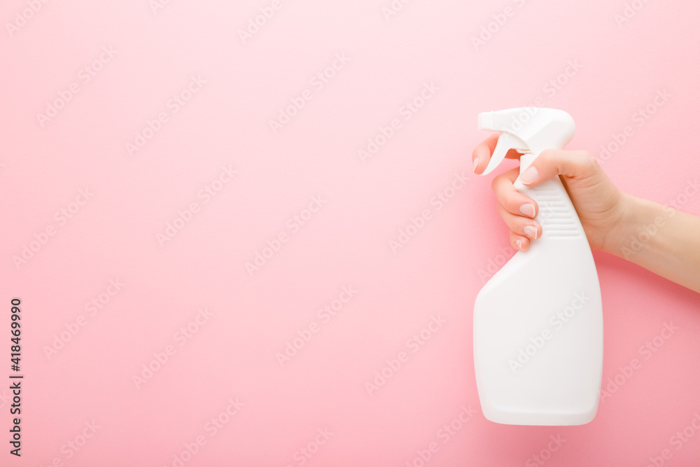 Young adult female hand holding white spray bottle. Detergent for different surfaces in kitchen, bathroom and other rooms. Closeup. Empty place for text or logo. Light pink background. Pastel color. - obrazy, fototapety, plakaty 