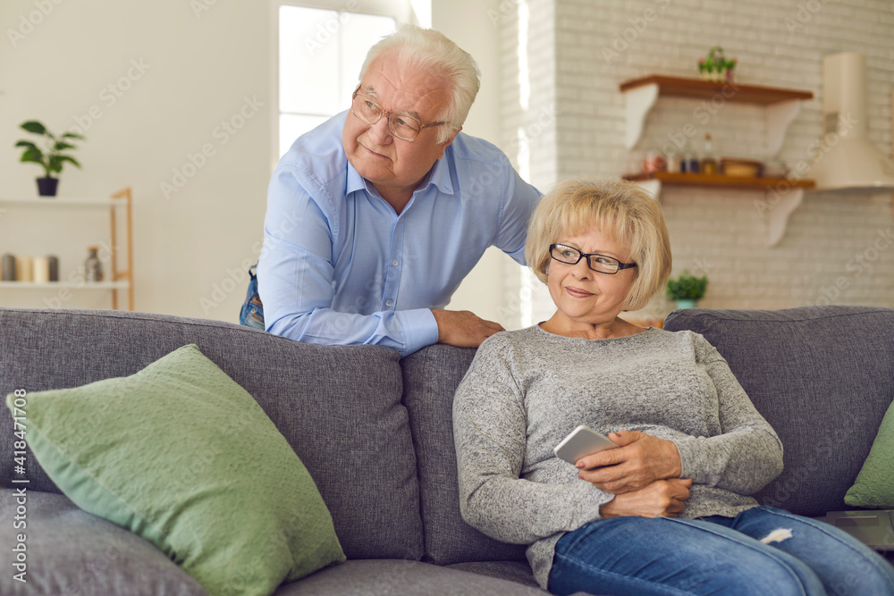 Happy old people enjoy free time in retirement. Senior couple staying at home together. Mature woman sitting on couch holding mobile phone, her husband leaning over, both looking away through window