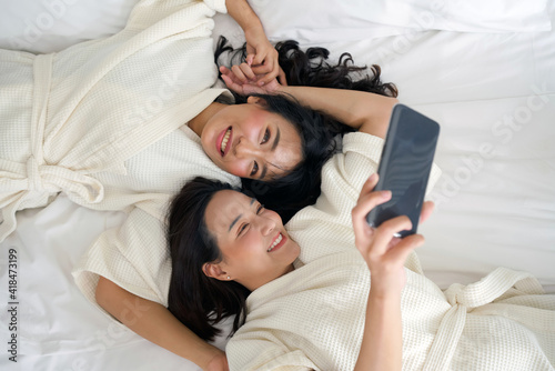 Two Asian women in bathrobes take a selfie with a mobile phone while lying in bed.