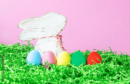 background for easter with a figurine of a rabbit with colored eggs in the grass on a pink background, place for text, symbol of light easter