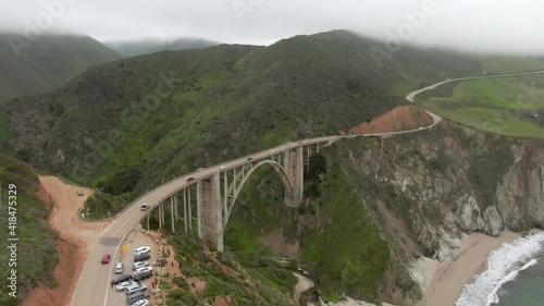 Aerial tilt down shot of vehicles on Bixby Creek Bridge by sea, drone flying forward towards green landscape during foggy weather - Big Sur, California