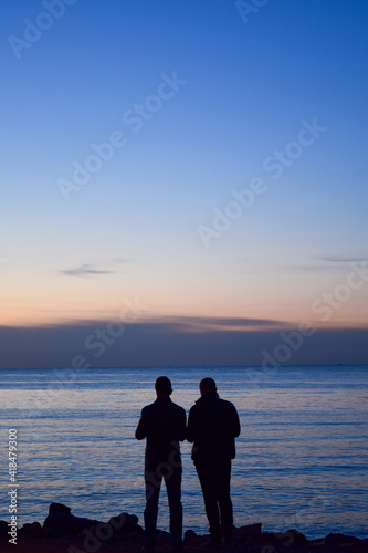 The silhouette of two young men standing on the cliff and taking photos for sunset 