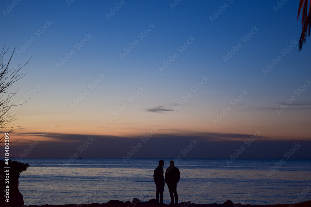 The silhouette of two young men standing on the cliff and taking photos for sunset  