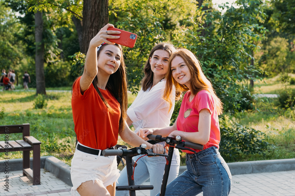 Obraz premium Three young girl friends on the electro scooters taking picture of themselves on cell phone at summer sunny day. Outdoor portrait of three friends girl riding electric kick scooter in the park