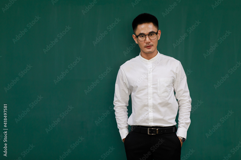 Portrait of young and handsome Asian man wearing eyeglasses and casual business clothes, white shirt and black trousers, standing pose with self-confidence with green background and copy space