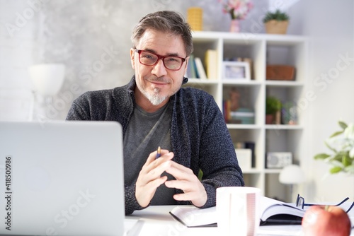 Bearded businessman working online with laptop computer at home sitting at desk. Home office, browsing internet, study room. Portrait of mature age, middle age, mid adult man in 50s.