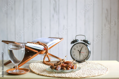 Iftar time cincpet. Kurma or dates fruit with glass of  water, holy Quran, alarm clock showing 6 o'clock and prayer beads on the table.  photo