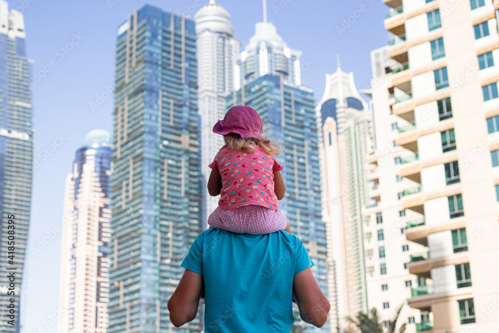 man holds a baby girl on his shoulders against the backdrop of skyscrapers.