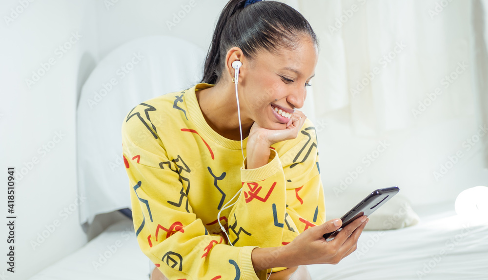 Cute black-haired brunette woman with yellow sweater talking on video call with smartphone and music headset while smiling and enjoying sharing and learning with communication from home Cute black-haired brunette woman with yellow sweater talking on video call with smartphone and music headset while smiling and enjoying sharing and learning with communication from home