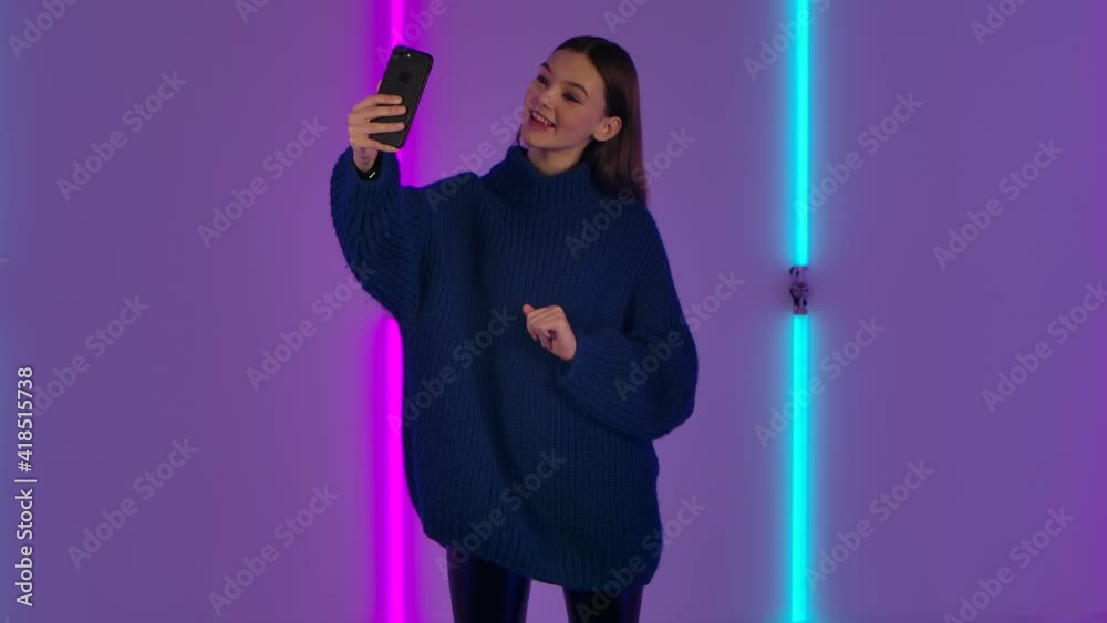 Portrait of fashionable model talking for video chat using mobile phone and rejoice. Young brunette woman with long hair in blue sweater poses against background of bright neon lights. Slow motion. Portrait of fashionable model talking for video chat using mobile phone and rejoice. Young brunette woman with long hair in blue sweater poses against background of bright neon lights. Slow motion.