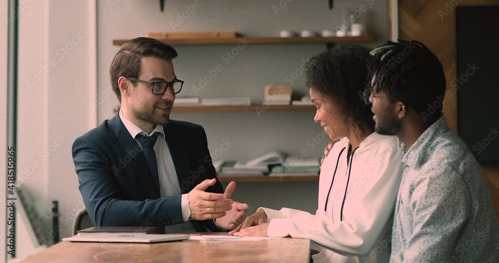 Smiling pleasant trusted young professional realtor or financial advisor in formal wear explaining offer issues to happy african american couple clients at meeting in office, shaking hands making deal