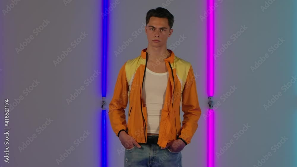 Portrait of stylish guy looking directly at the camera. Male fashion model in yellow jacket poses in dark studio against the backdrop of bright neon lights. Slow motion. Portrait of stylish guy looking directly at the camera. Male fashion model in yellow jacket poses in dark studio against the backdrop of bright neon lights. Slow motion.