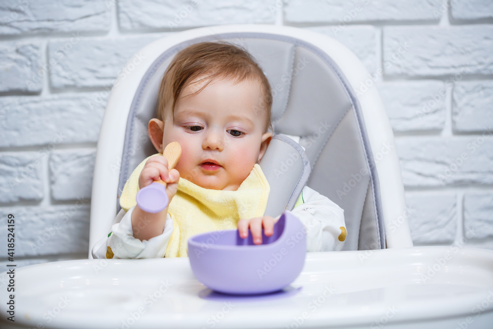 A small child sits on a highchair and eats food from a plate with a spoon. Baby silicone utensils for feeding babies A small child sits on a highchair and eats food from a plate with a spoon. Baby silicone utensils for feeding babies
