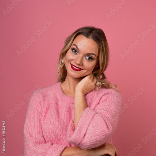 Young attractive woman in a warm pink sweater on a pink background, blonde with curly hair and red lipstick