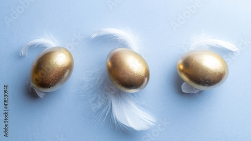 Easter egg. Happy Easter decoration: Golden eggs with white feathers on pastel blue background. Traditional decoration in sun light. Top view.