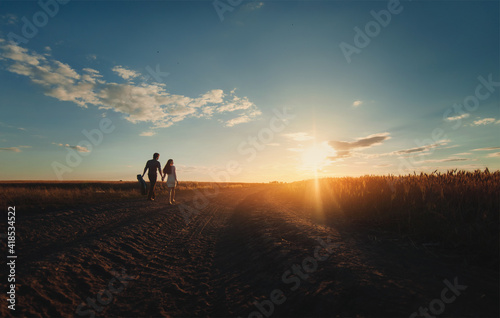 A young newlyweds couple in love is walking holding hands to meet the sunset.Silhouette of a guy with a guitar who leads his girlfriend along road between wheat fields.Country style.View from the back