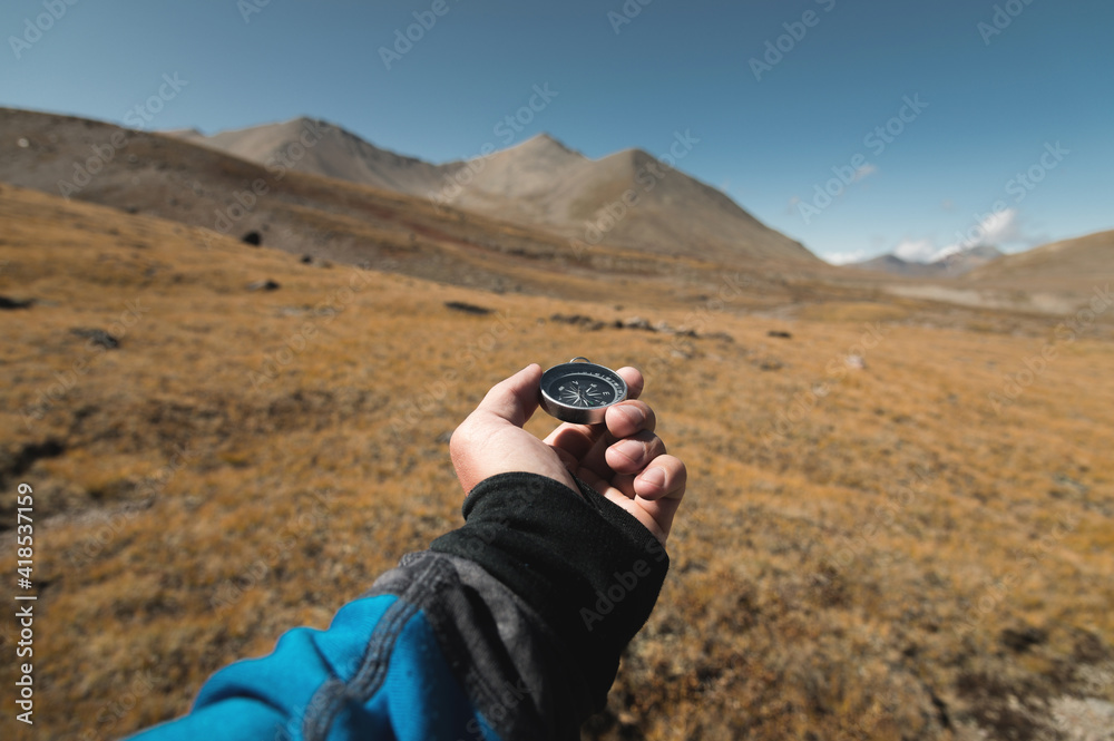 First-person view of a male traveler's hand holding a magnetic compass against the backdrop of a mountainous area. Orientation and finding your way