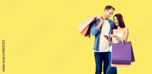 Love, holiday sales, shop, retail, consumer concept - happy smiling couple with shopping bags, and cellphone, standing close to each other. Studio portrait of young man and woman, isolated over yellow