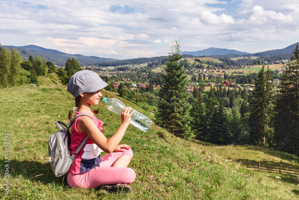 Little girl in a T-shirt with a backpack on a background of mountain landscape. The child drinks water from a bottle sitting on a hill and looking into the valley
