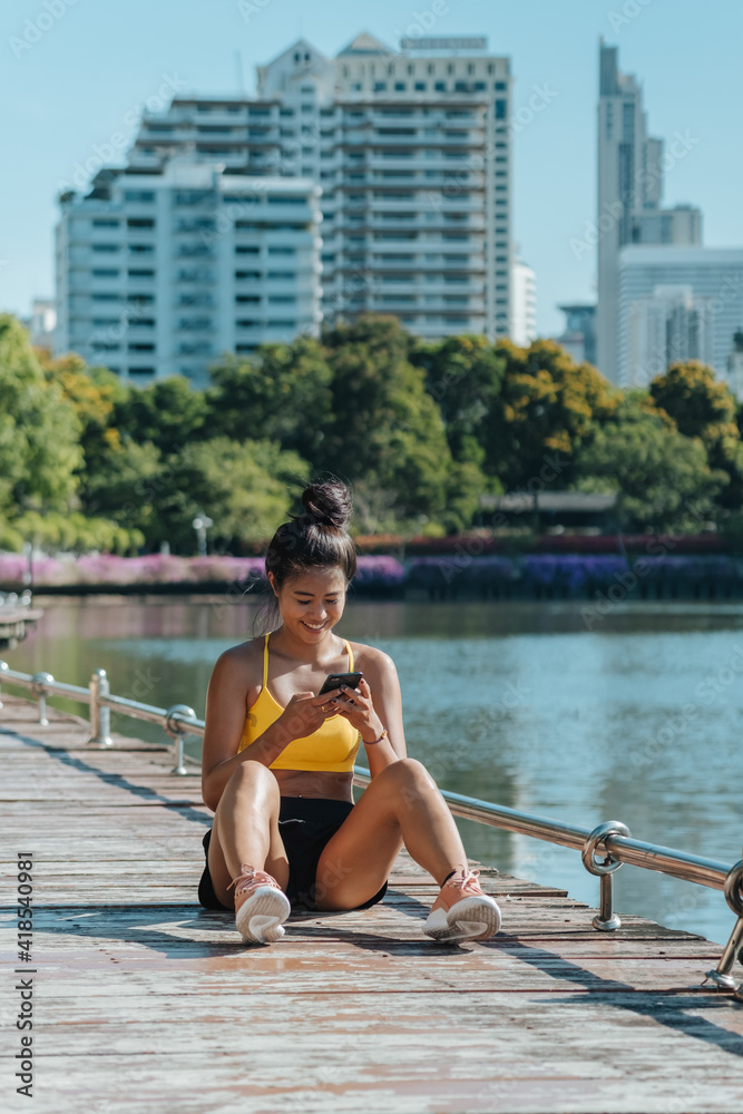 Smiling woman in in activewear resting and using mobile phone fitness app for tracking performance after workout in the park with river and city buildings in the background Smiling woman in in activewear resting and using mobile phone fitness app for tracking performance after workout in the park with river and city buildings in the background