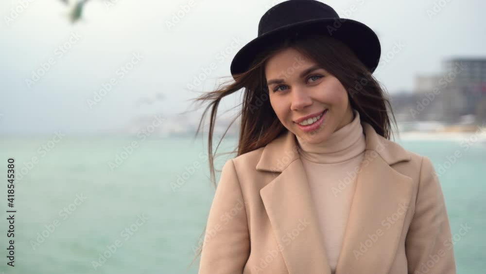 Close-up portrait, a young Caucasian girl stands on the pier dressed in a coat and hat near the ocean or sea and looks into the camera. Copy space. confidential smile. Close-up portrait, a young Caucasian girl stands on the pier dressed in a coat and hat near the ocean or sea and looks into the camera. Copy space. confidential smile.
