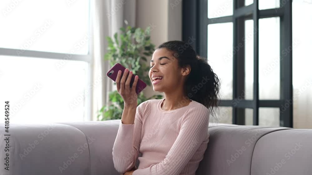Young mixed-race ethnic woman or female student with afro hairstyle wearing casual clothes, sitting on the couch at home and holding a mobile phone, talking, gesturing, recording voice message Young mixed-race ethnic woman or female student with afro hairstyle wearing casual clothes, sitting on the couch at home and holding a mobile phone, talking, gesturing, recording voice message