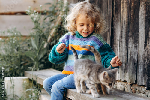 Portrait of a beautiful and happy curly girl in a knitted sweater playing with a kitten near a wooden wall