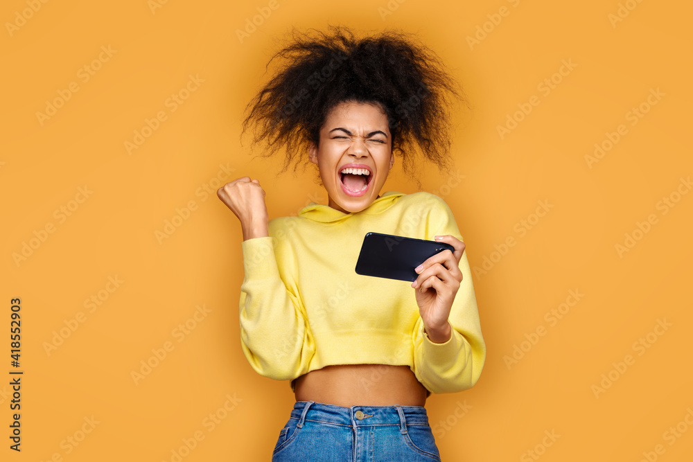 Happy girl clenched fist in victory gesture, victory in the game. Photo of african american girl using smartphone on yellow background Happy girl clenched fist in victory gesture, victory in the game. Photo of african american girl using smartphone on yellow background