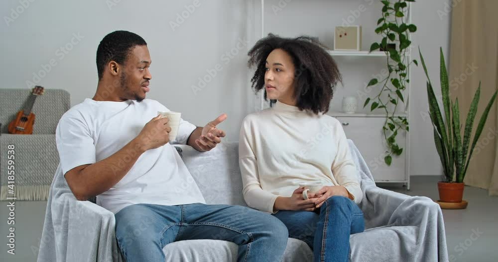 Afro american couple family handsome man and curly attractive girl wearing casual clothes at home discussing sitting on couch drinking tea hot coffee in morning enjoying weekend break chatting talking Afro american couple family handsome man and curly attractive girl wearing casual clothes at home discussing sitting on couch drinking tea hot coffee in morning enjoying weekend break chatting talking