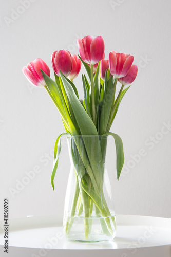 A bouquet of pink tulips in a glass vase on a white metallic table in front of a light grey wall.