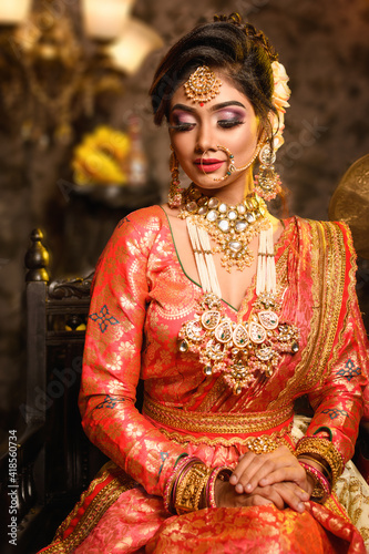 Leinwand Poster Magnificent young Indian bride in luxurious bridal costume with makeup and heavy jewellery is sitting in a chair in with classic vintage interior in studio lighting