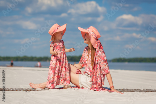 A little girl with her mother in matching beautiful sundresses plays in the sand on the beach. Stylish family look photo