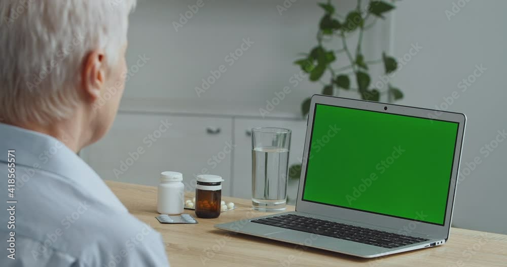 Close-up view from behind gray-haired unrecognizable old woman grandmother sick patient looks at green screen modern laptop sits at table with bottle of pills medicine uses online technologies at home Close-up view from behind gray-haired unrecognizable old woman grandmother sick patient looks at green screen modern laptop sits at table with bottle of pills medicine uses online technologies at home