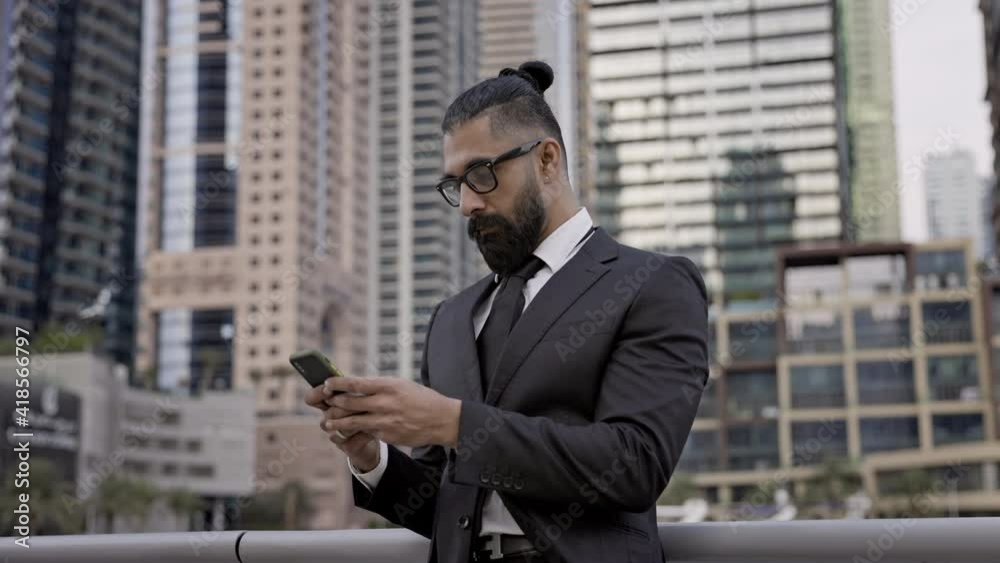 Stylish Young Man In Business Suit On Background Of The City. Tall Buildings Dubai. He Holds Mobile Phone And Looks At It. Stylish Young Man In Business Suit On Background Of The City. Tall Buildings Dubai. He Holds Mobile Phone And Looks At It.