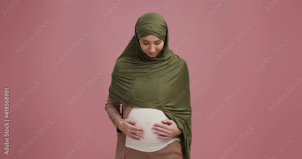 Young muslim pregnant lady in hijab caressing her big belly, enjoying her pregnancy, pink background Young muslim pregnant lady in hijab caressing her big belly, enjoying her pregnancy, pink background