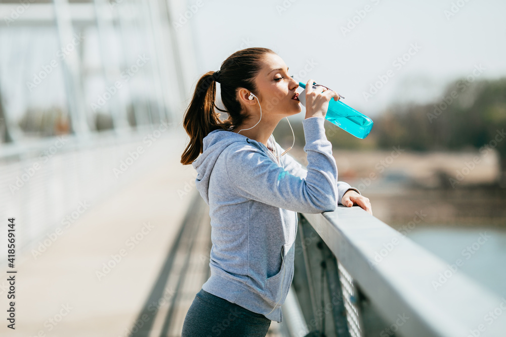 Beautiful young and fit woman in good shape relaxing after running and jogging alone on city bridge street. She wears sporty earbuds and listens to music.