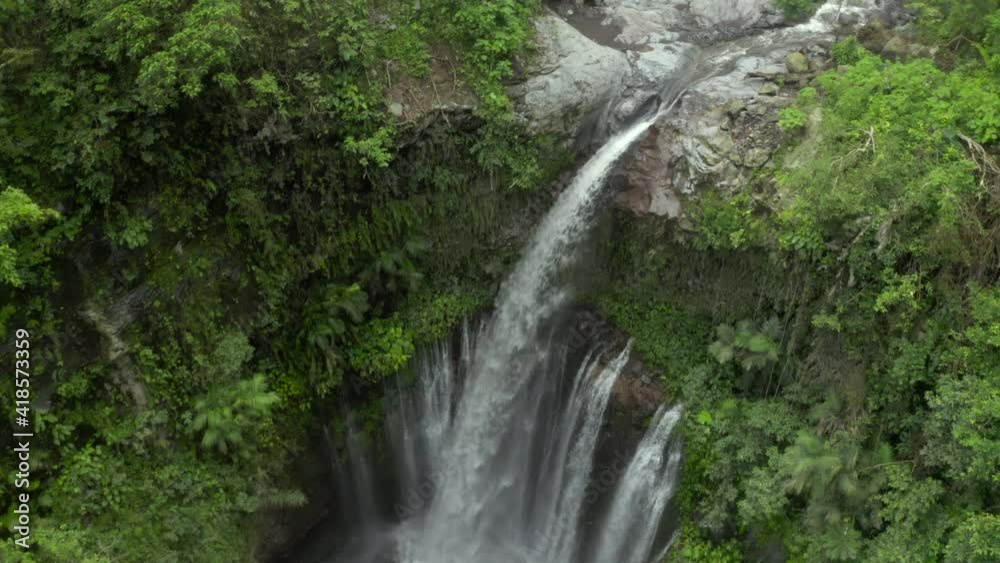 Aerial tilt down to a plunge basin of a rushing waterfall surrounded by jagged cliff walls covered in dense tropical foliage with cool mist clinging to the treetops - Nusa Penida & Lombok, Bali Aerial tilt down to a plunge basin of a rushing waterfall surrounded by jagged cliff walls covered in dense tropical foliage with cool mist clinging to the treetops - Nusa Penida & Lombok, Bali