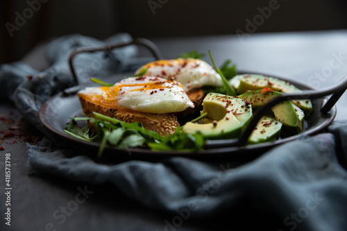 Delicious and delicious breakfast with poached egg on a slice of fried bread, avocado, arugula, cherry tomatoes and spices. Served in a cast-iron skillet, served with textiles on a black background . 