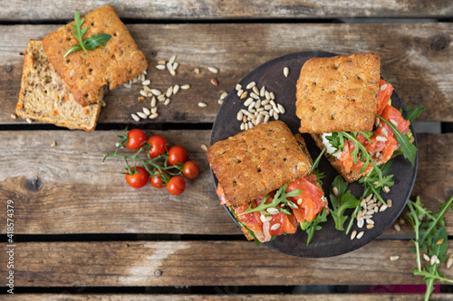  A delicious snack for lunch. Delicious sandwich with lightly salted salmon. With a lush bun, with arugula greens and sunflower seeds, with cherry tomatoes and avocado on the wood background.