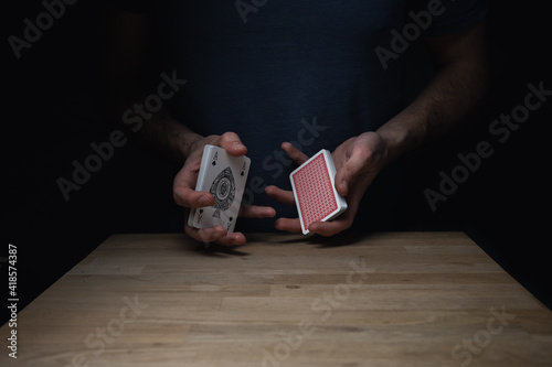 Man in blue shirt against dark background holding a split deck of red cards at a wooden table showing the ace of spades. Games cards playing poker gambling.